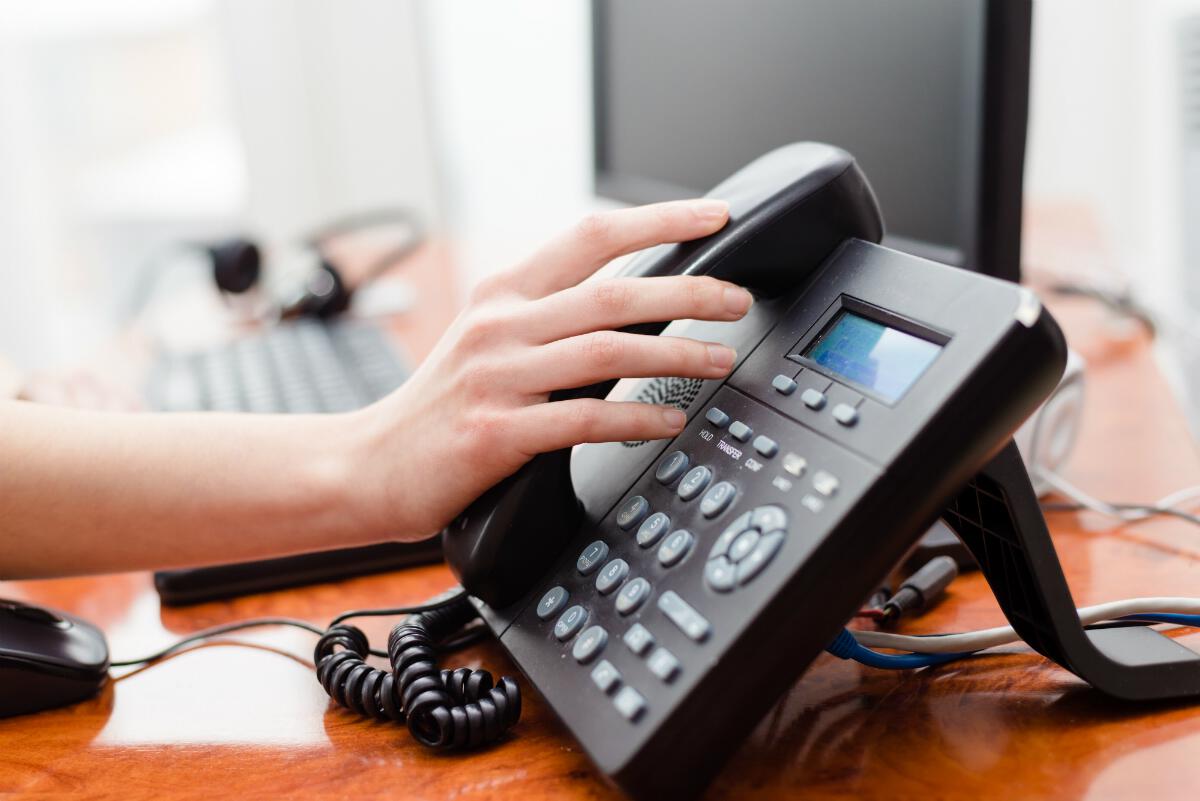 A hand resting on a phone receiver still on the base, with other office equipment out of focus on the same desk