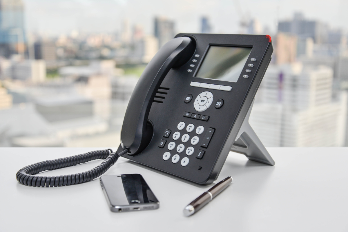 A business phone on a desk, with a blurred city view in the background and a smart phone and pen in the foreground
