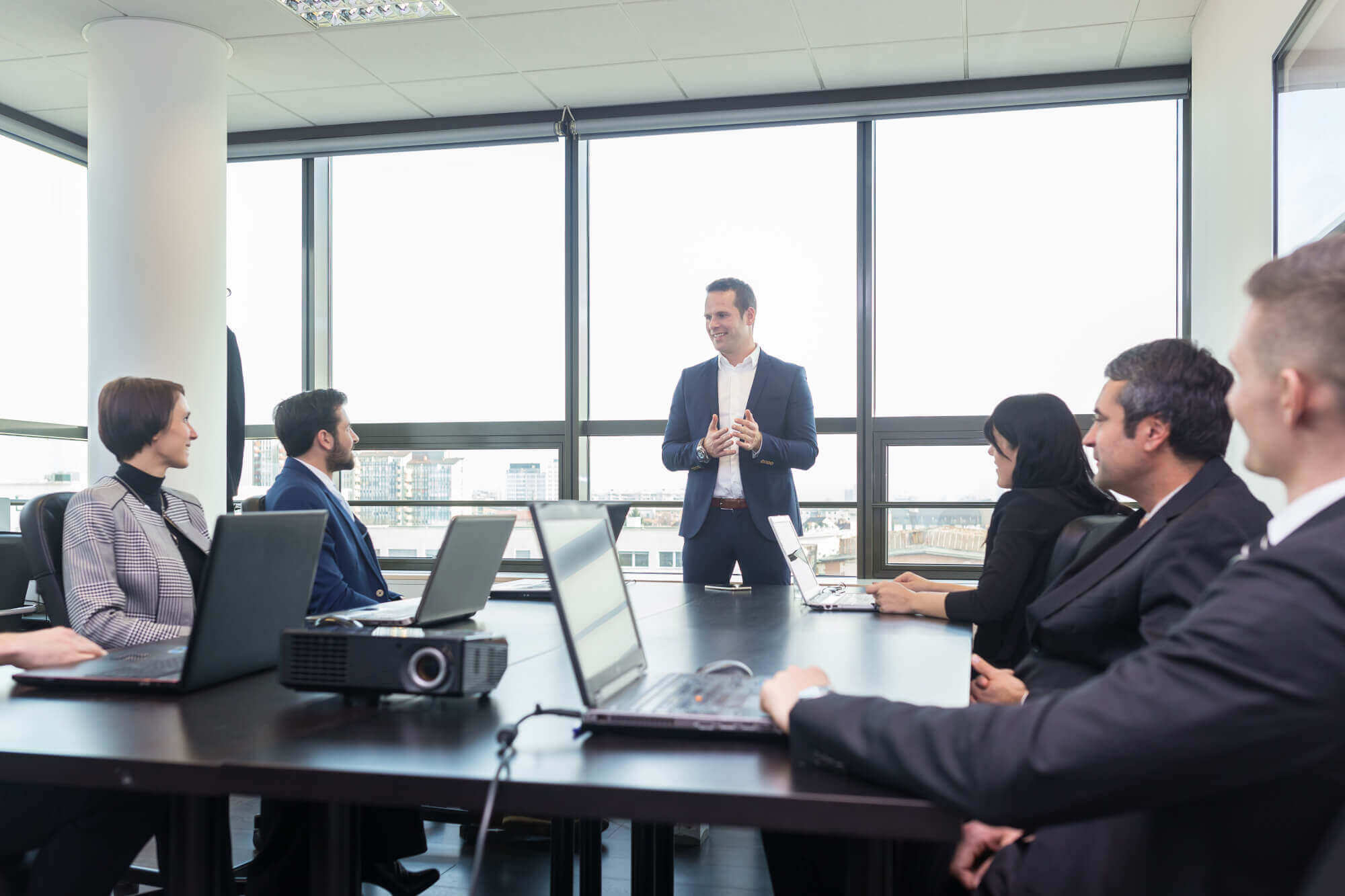 Businessman standing at head of table and speaking to colleagues using laptops